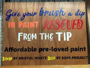 A wooden sign that reads in paint: give your brush a dip in paint rescued from the tip. Affordable pre-loved paint. Saved by Bristol Waste. Sold by Sofa Project.
