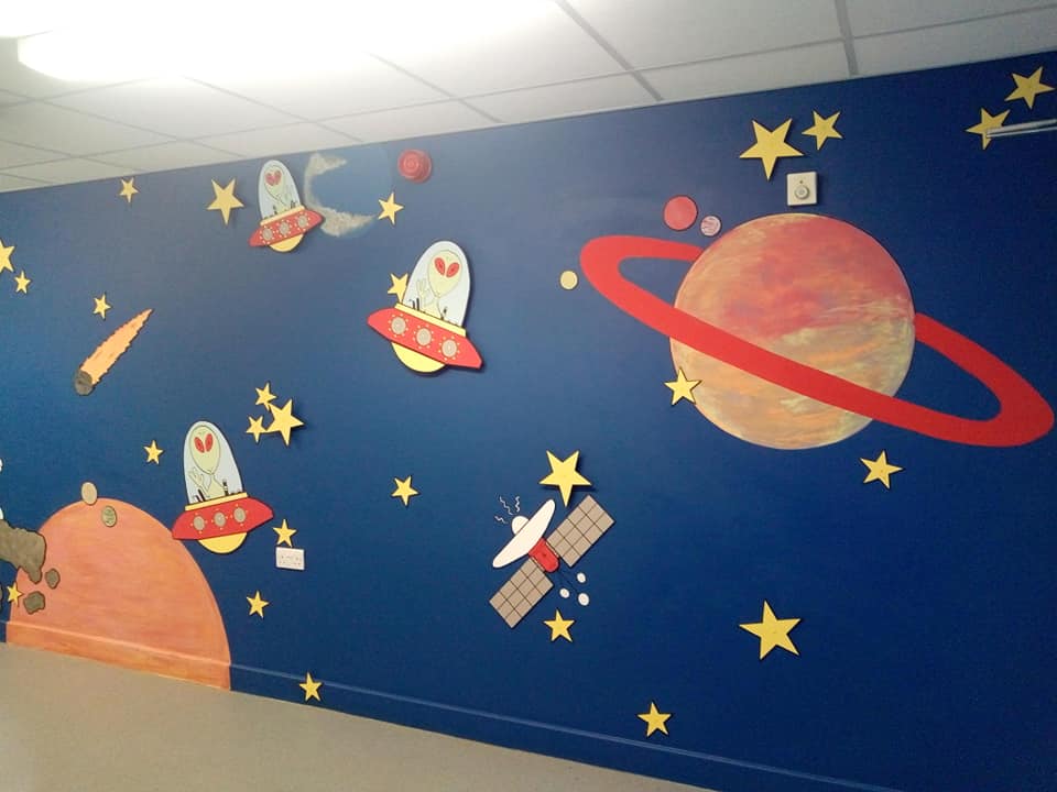 A painted mural at a school in Bradford using paint from Community RePaint Bradford'sIt shows carefully cut out space shapes from MDF on a background of dark blue silk emulsion paint. Planets have also been painted on the dark blue background in red and orange recycled matt emulsion.