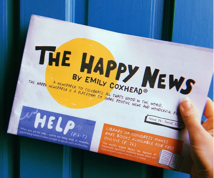 A hand holding onto a community newspaper that reads: The Happy News by Emily Coxhead. A newspaper to celebrate all that's good in the worls. The happy newspaper is a platform to share positive news and wonderful people.