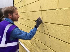 A volunteer painting the community mural at the Bradford Interchange. The volunteer is painting the outline of a notable building on a yellow paint panel.