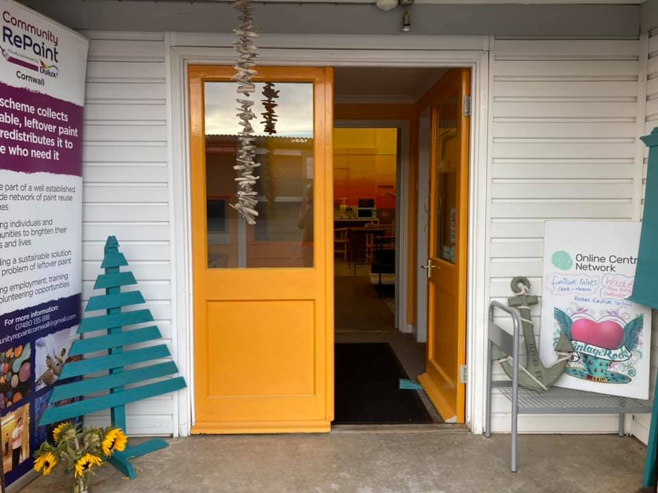 A photo of the newly painted door and entrance to the Community RePaint Cornwall scheme. The front door has been painted in a warm yellow and there is a upcycled crate that has been converted into the shape of a tree that has been painted green.