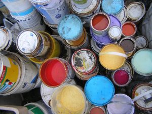 A photo looking down onto a variety of cheap and recycled paint. Some of the paint containers have their lids removed to show you the range of colours which include reds, yellows and blues.